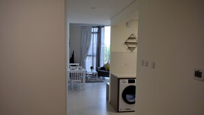 Ett modernt kök med en inbyggd tvättmaskin, en matplats med ett vitt bord och stolar samt ett vardagsrum med en soffa och ett fönster med gardiner.