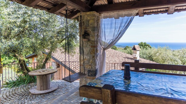 Une table et un banc en bois, placés sous une terrasse couverte, avec un pilier en pierre et une vue sur la mer.