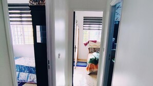 Un couloir étroit, dont le sol est recouvert de carreaux, mène à une chambre à coucher équipée d’un lit et d’une fenêtre avec des stores.
