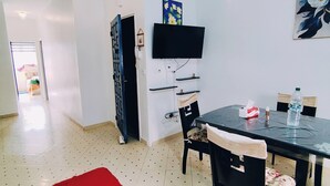 Un coin repas comprenant une table, des chaises, un tapis rouge, un téléviseur fixé au mur et une porte donnant sur une autre pièce.
