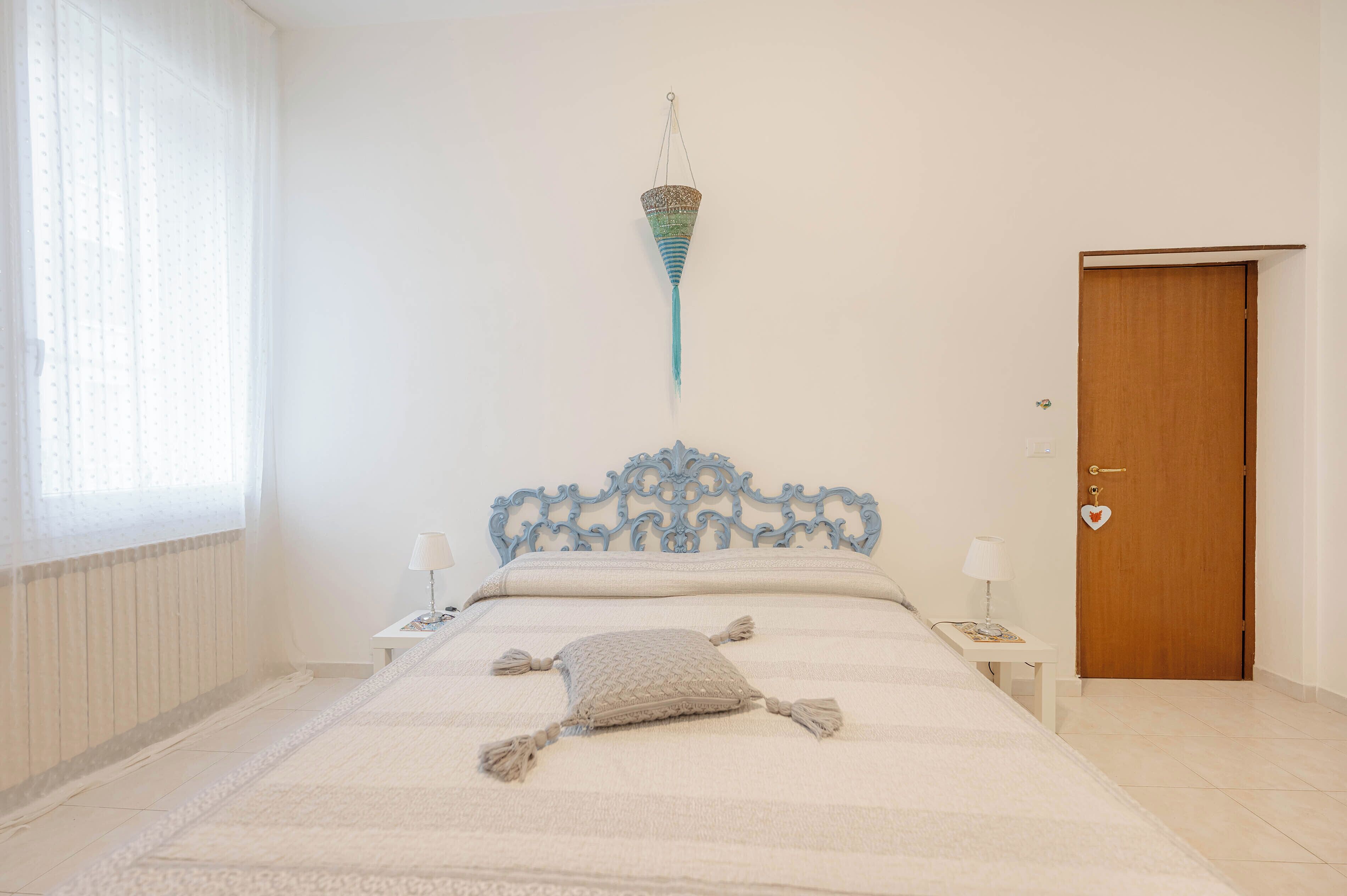 A bedroom with a bed, two bedside tables, a hanging light fixture, a wooden door, and a window with sheer curtains.