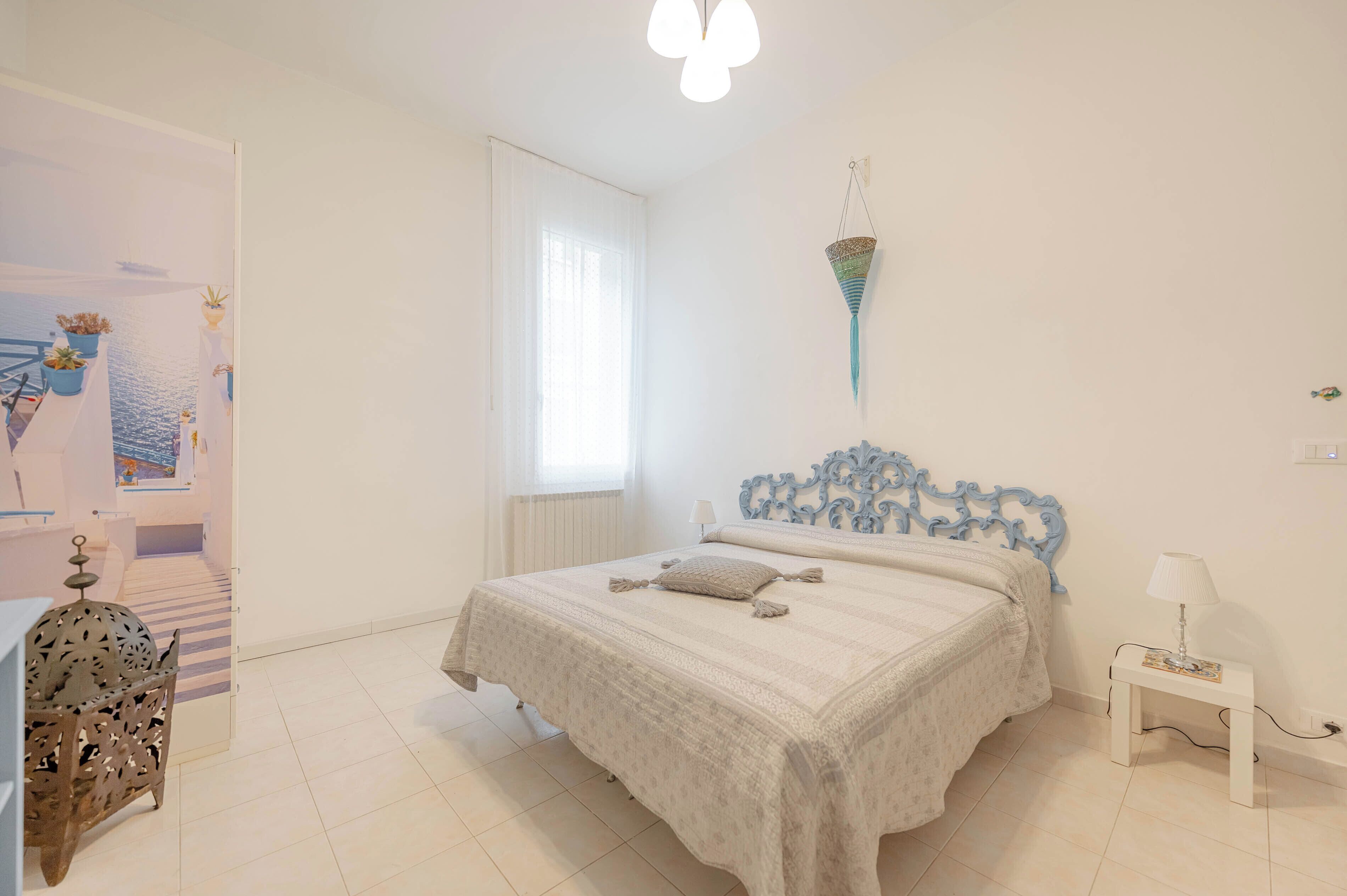 A bedroom with a bed, a bedside table with a lamp, a decorative wall hanging, and a view of a balcony with potted plants.