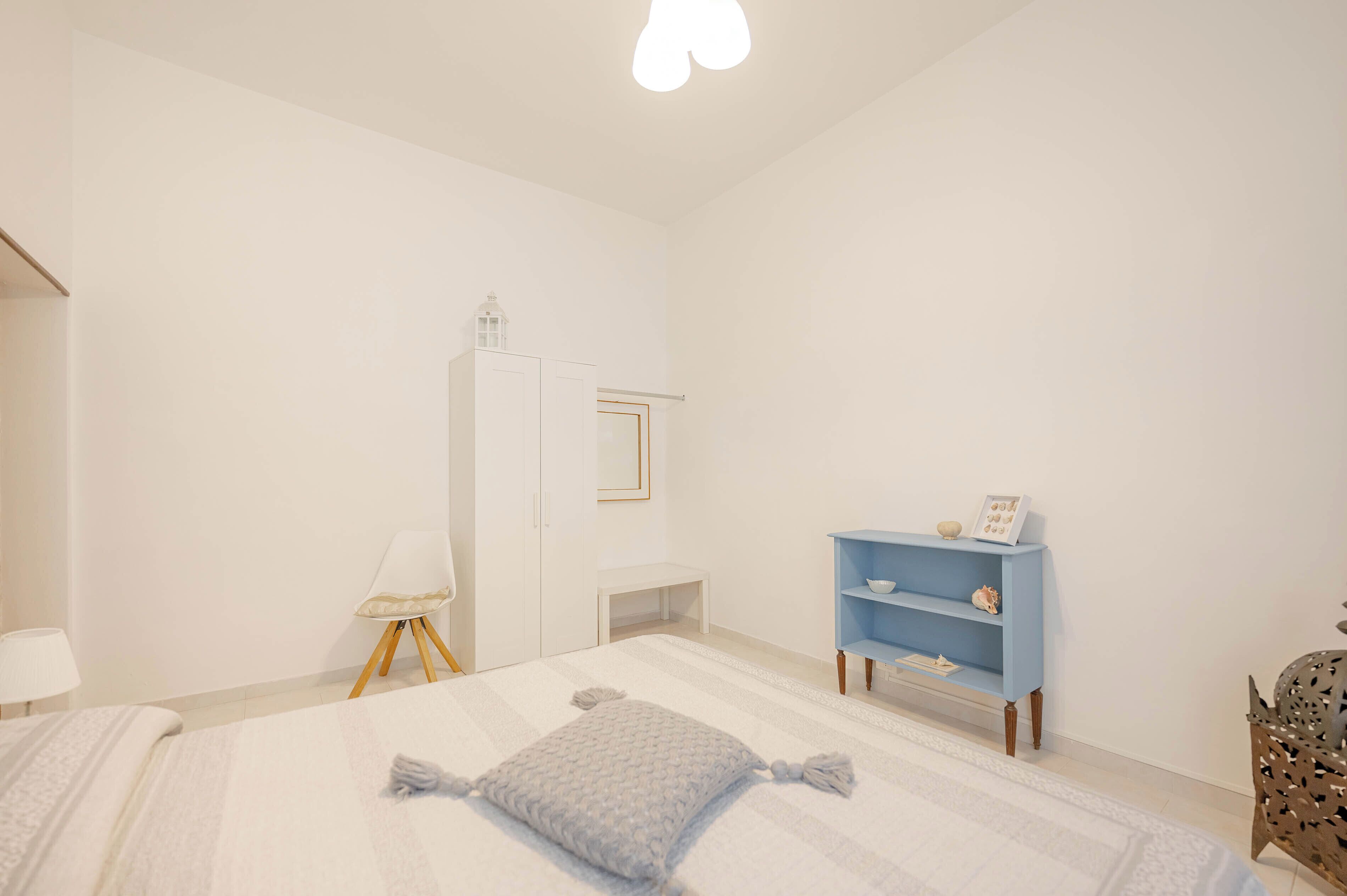 A bedroom with a bed, a white wardrobe, a blue shelf, a chair, and a bedside table.