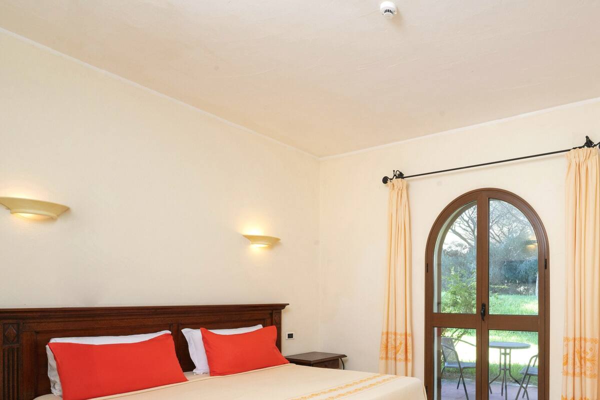 Une chambre avec un grand lit, deux appliques murales, une tête de lit en bois, une table de chevet, une fenêtre avec des rideaux et une vue sur la verdure à l’extérieur.