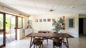 En rummelig stue med et stort træbord, fire stole, en potteplante og en brandslukker på væggen.