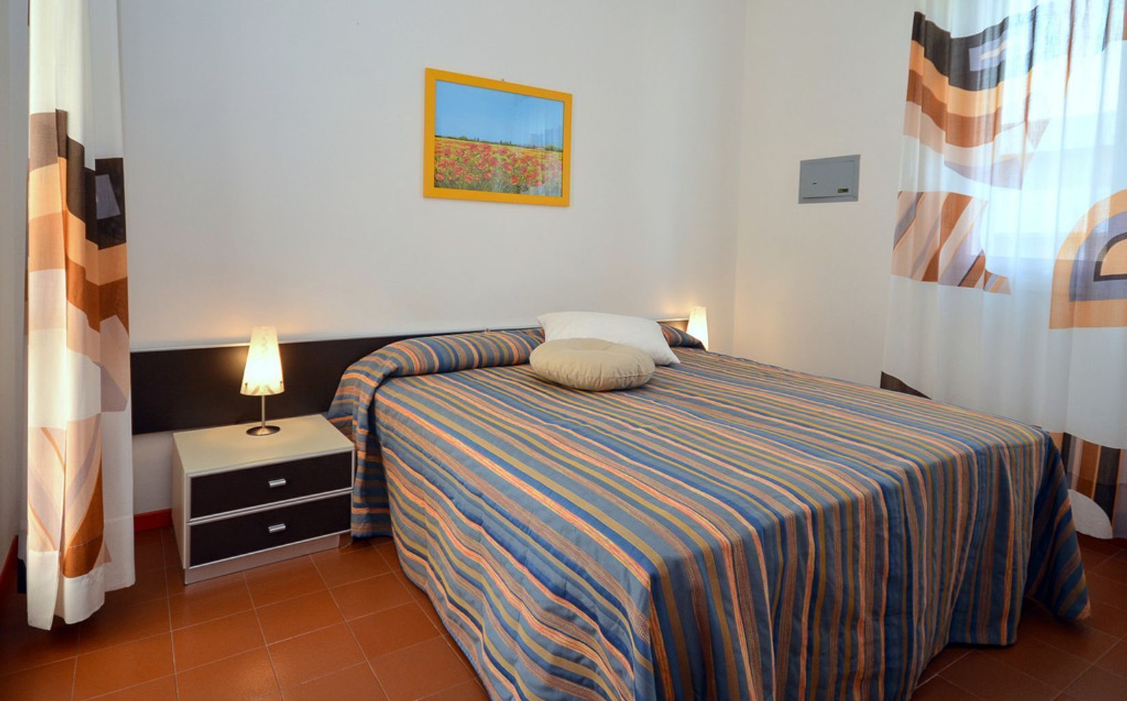 Un dormitorio con cama, mesitas de noche, un cuadro en la pared y cortinas.