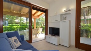 A living room with a blue sofa, a TV on a white cabinet, and a sliding glass door leading to an outdoor patio with chairs and greenery.