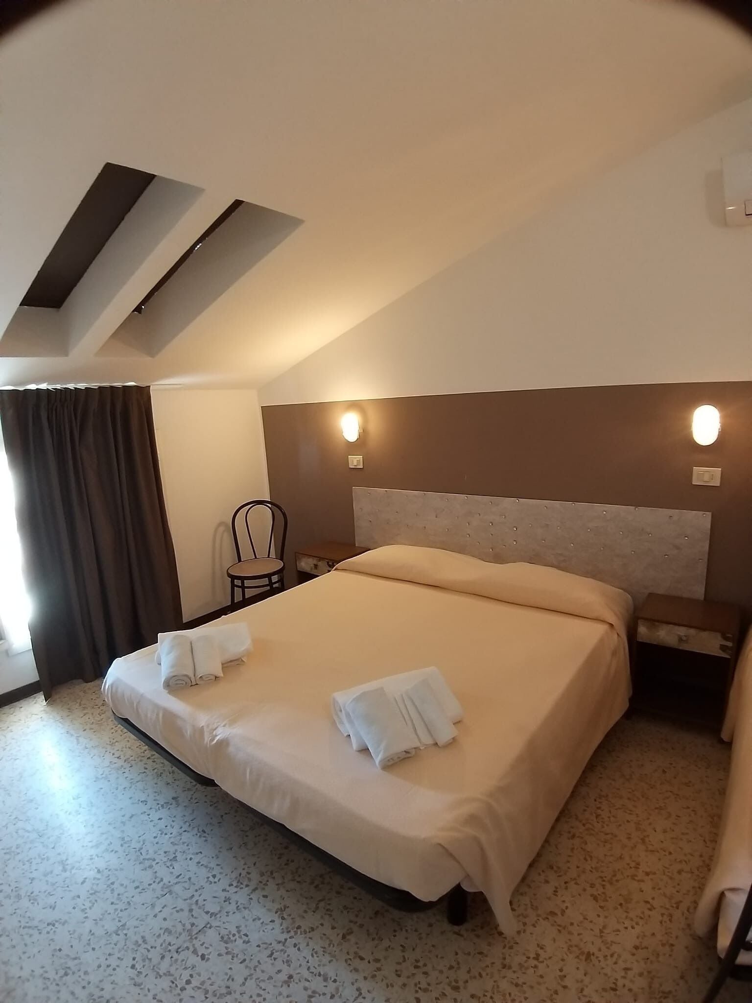 A hotel room with a large bed, two wall-mounted lights, a chair, and a desk.
