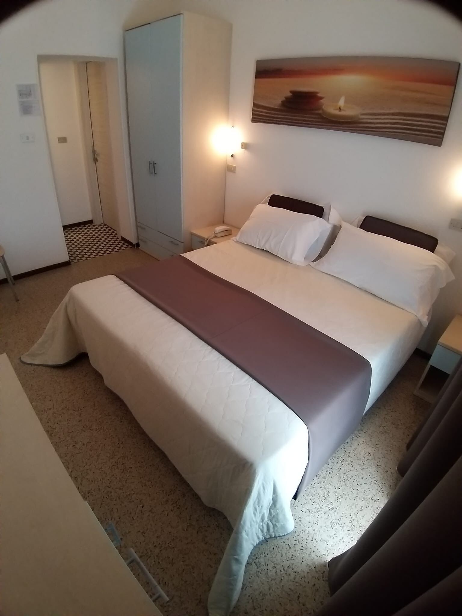 A hotel room with a bed, a nightstand, a lamp, a wardrobe, and a painting on the wall.