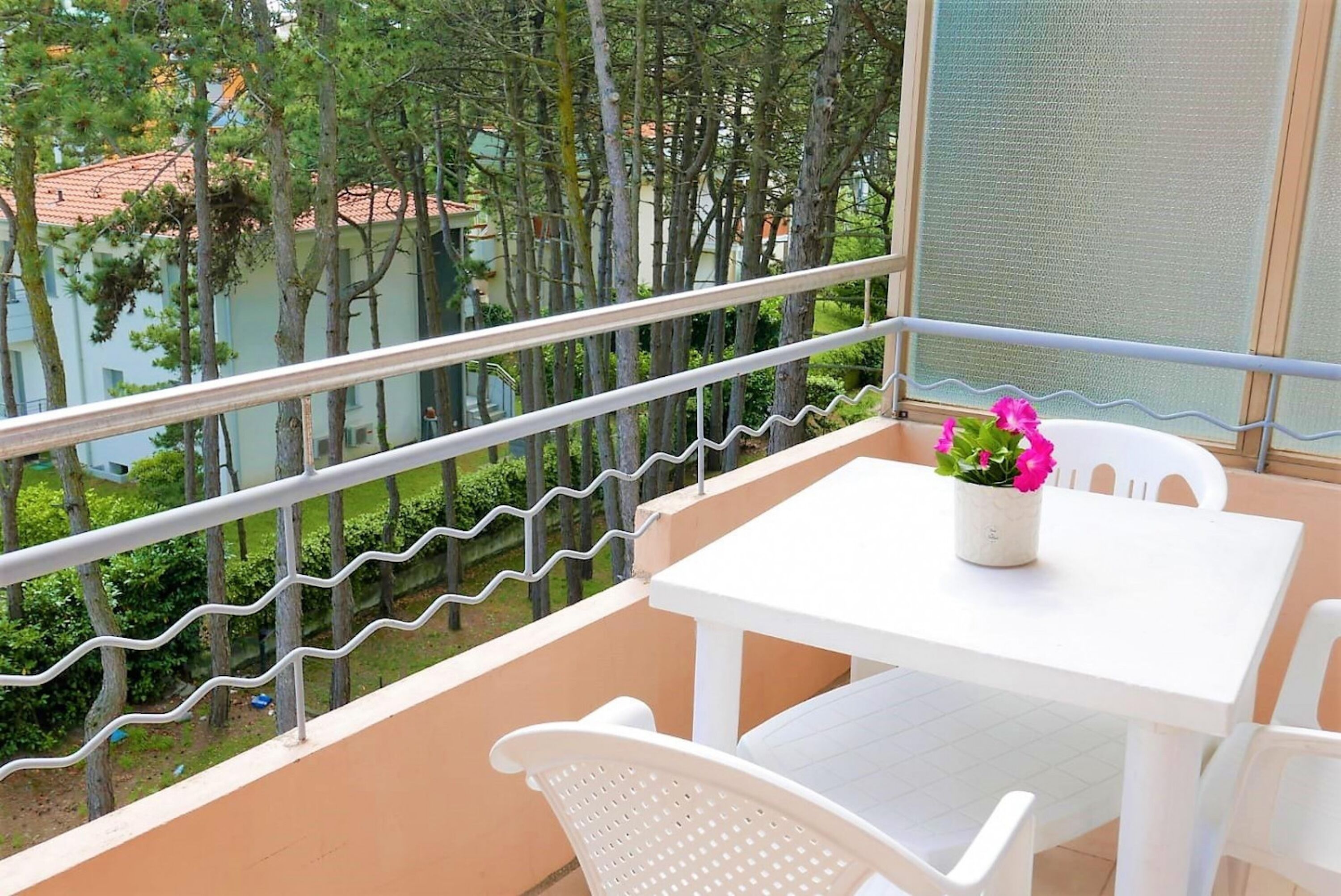 Een balkon met een witte tafel en stoelen, een bloempot met roze bloemen en uitzicht op bomen en gebouwen.