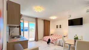 Ein Hotelzimmer mit Küchenzeile, einem Bett mit floralem Überwurf, einem an der Wand montierten Fernseher und einem Balkon mit Blick auf Gebäude.