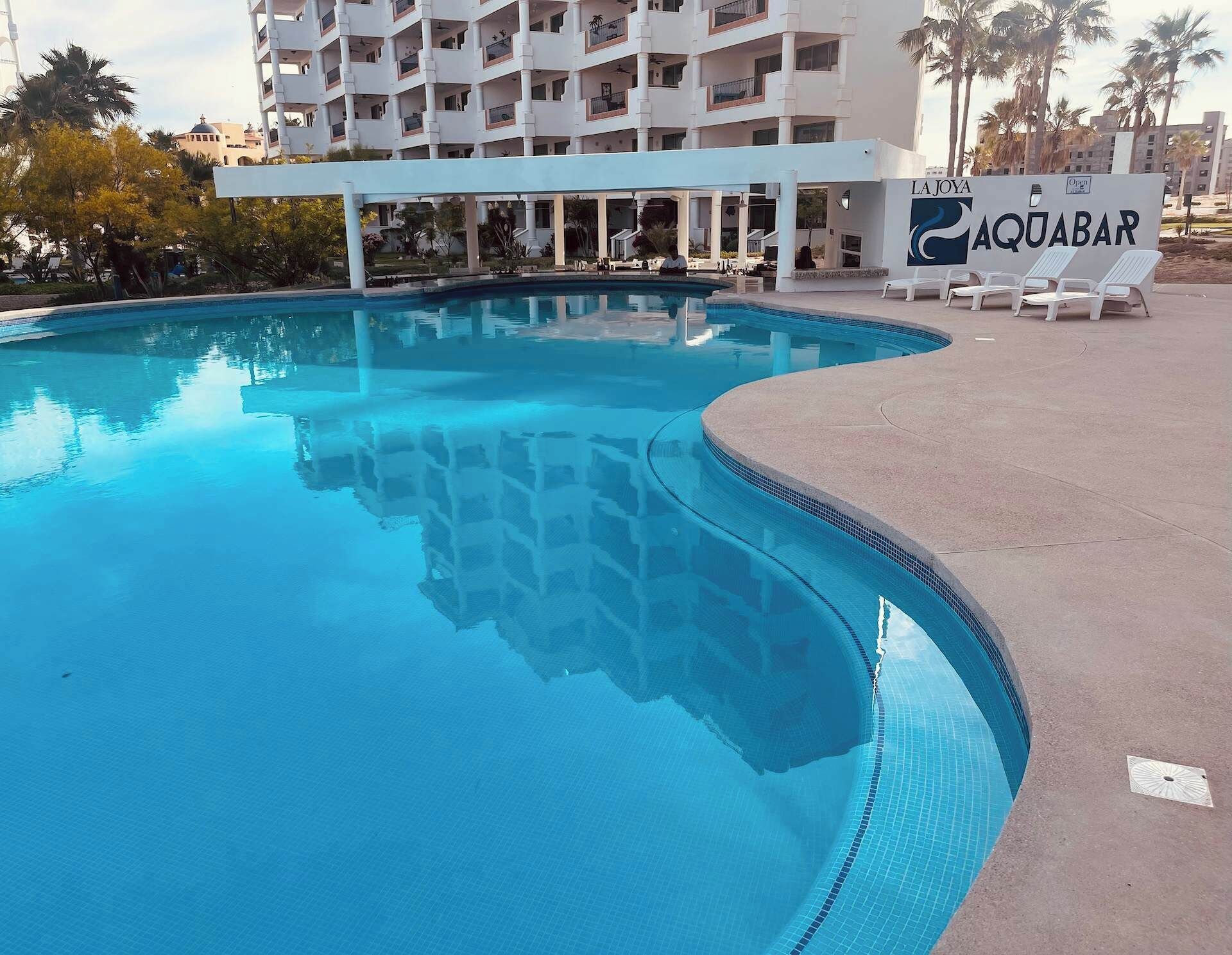 Une piscine à bordures courbes, entourée de chaises longues et d’un panneau indiquant « LA JOYA AQUABAR ».
