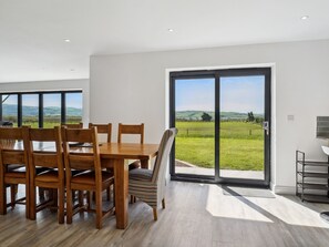 Ein Essbereich mit Holztisch und -stühlen, großen Schiebeglasfenstern zu einem grünen Feld und einer modernen Küchenzeile.