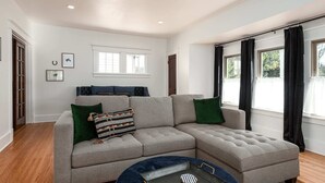 Ein modernes Wohnzimmer mit Ecksofa, rundem Couchtisch und großem Fenster mit Vorhängen.