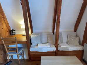 Une chambre de style « loft » avec des poutres en bois apparentes, une chaise en bois, une petite table avec une lampe et un canapé avec des coussins.
