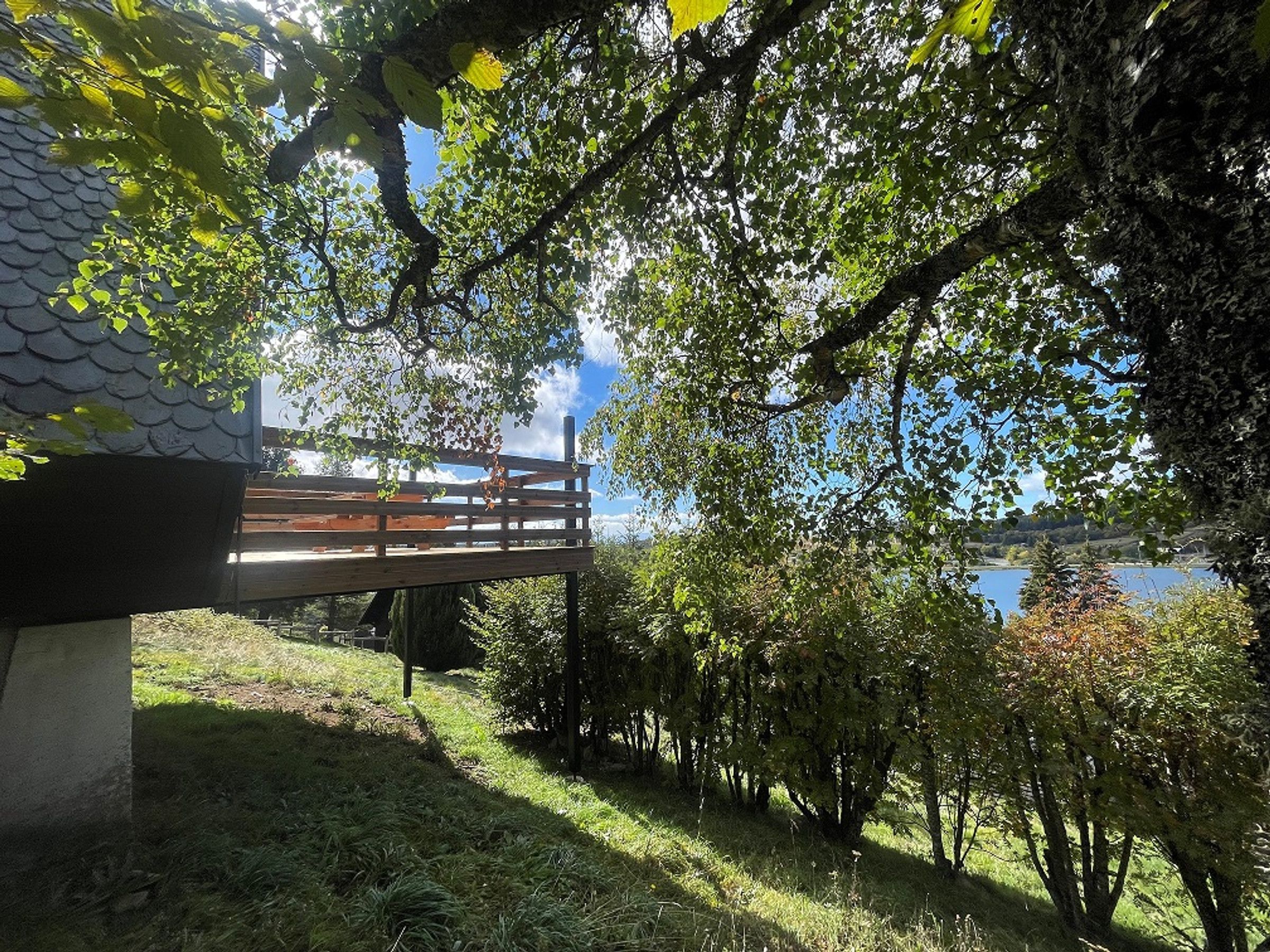 Une terrasse en bois surplombant une étendue d’eau, entourée d’arbres et d’un bâtiment doté d’un toit en tuiles.
