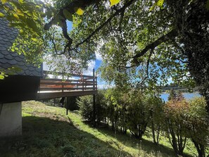 Eine Holzterrasse mit Blick auf ein Gewässer, umgeben von Bäumen und einem Gebäude mit Ziegeldach.