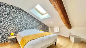 A bedroom with a sloped ceiling, a skylight, a bed with a yellow blanket, a small table with a lamp, and a patterned wallpaper. - Charming 4-Bedroom Cottage with Modern Comfort (Haroué)