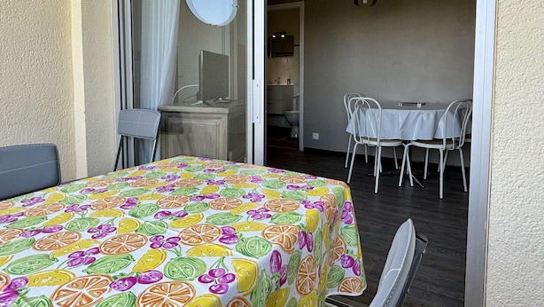 Un balcón con una mesa y sillas, una puerta corrediza de vidrio y una pequeña cocina con un lavamanos y un inodoro visibles a través de la puerta.