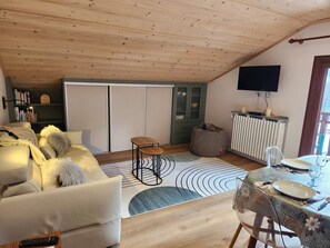 Un salon chaleureux avec un plafond haut, un canapé, une petite table, un fauteuil poire et un coin repas avec une table mise pour deux personnes.