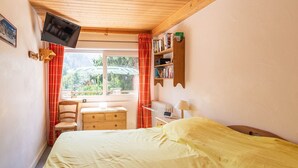 Une chambre à coucher comprenant un lit, un bureau, une chaise, un téléviseur, une fenêtre avec des rideaux et une bibliothèque.