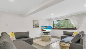 Un salon moderne avec des canapés gris, un téléviseur à écran plat, une petite table d’appoint en osier et une grande fenêtre donnant sur un paysage verdoyant.