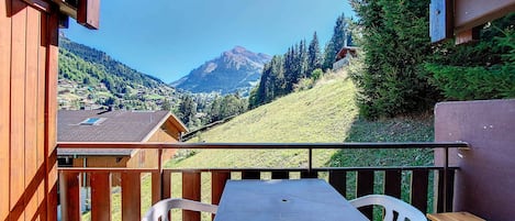 A wooden balcony with a table and chairs, overlooking a mountainous landscape with houses and trees.