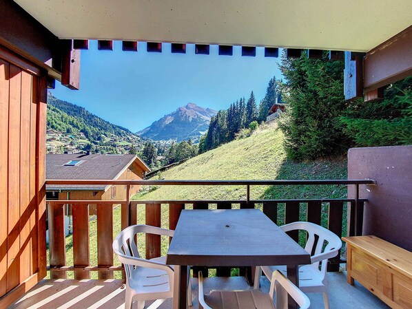A wooden balcony with a table and chairs, overlooking a mountainous landscape with houses and trees.