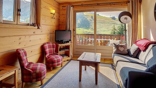 Interior acogedor de una cabaña con paredes de madera, un sofá, dos sillones a cuadros, un televisor y un ventanal con vistas a la montaña.