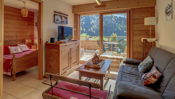 Ein gemütliches Zimmer mit Bett, Sofa, Fernseher und Tisch. Ein großes Fenster bietet Blick auf Bäume und Berge.