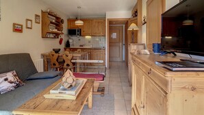 Ein gemütliches Wohnzimmer mit einem Holztisch, einer Couch und einem Fernseher. Im Hintergrund ist eine Küche mit Holzschränken und einem großen Fenster zu sehen.