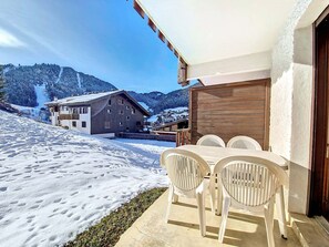 A snow-covered outdoor area with a table and chairs, a wooden building in the background, and a mountainous landscape.