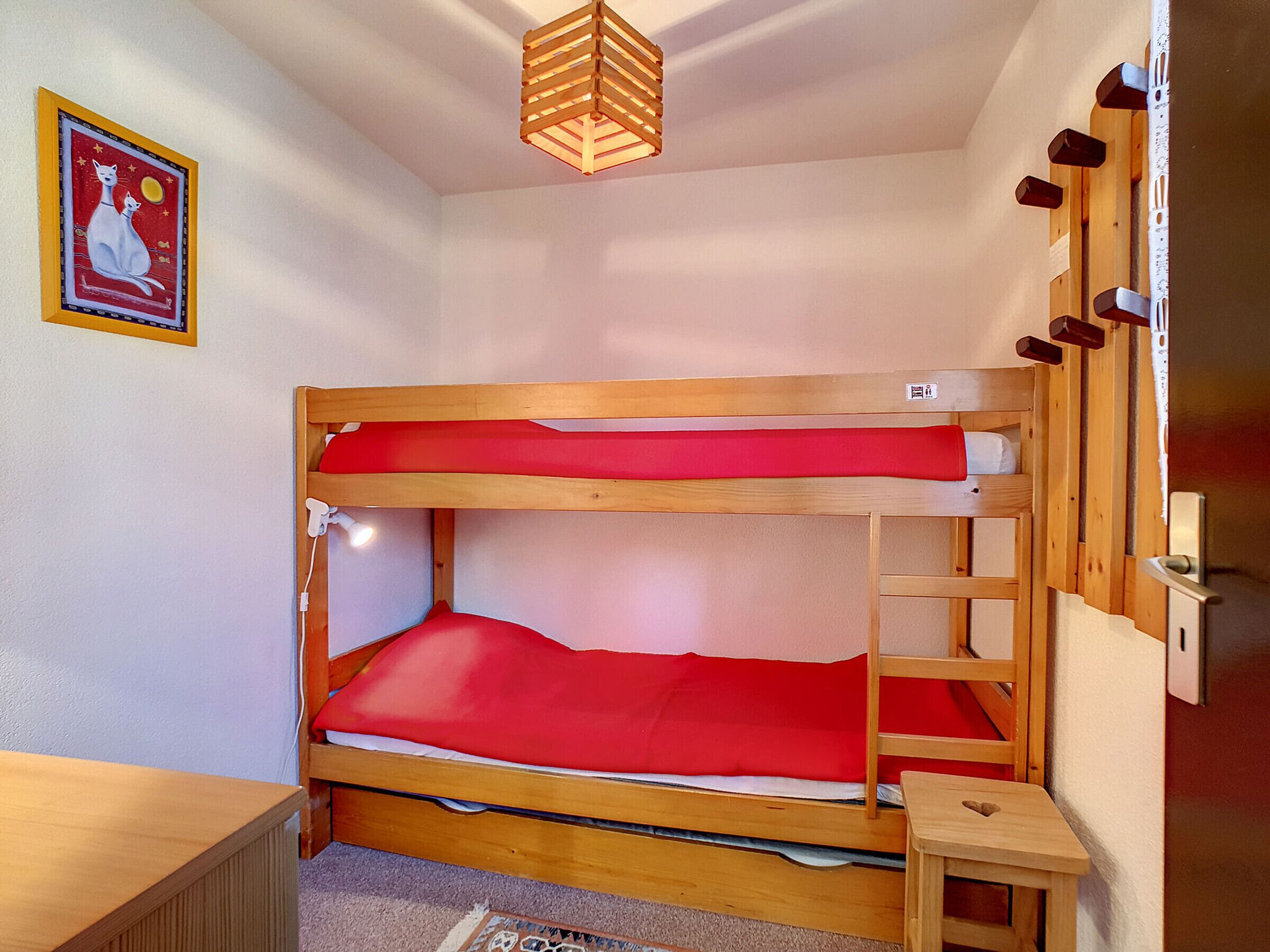 Habitación con literas, un colchón rojo, una pequeña mesa de madera, un estante en la pared y una lámpara del techo.