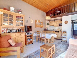 A cozy living room with a sofa, a coffee table, and a TV. There is a kitchen area with wooden cabinets and a dining table.