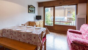 Ein Essbereich mit einem Tisch für vier Personen, einer Bank, einer Couch, einem Fernseher und einem Fenster mit Blick auf ein Gebäude und Bäume.