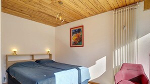 Un dormitorio con techo de madera, una cama con edredón azul, una silla roja y un cuadro en la pared.
