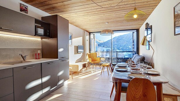 Una cocina moderna con un gran ventanal, techo de madera y una zona de comedor con una mesa dispuesta para cuatro.