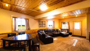 A cozy living room with a wooden ceiling, a leather sofa, a dining table with chairs, and a large window with curtains. - Warm River Butte Lodge (Ashton)