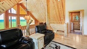 A spacious loft-style room with a high ceiling, wooden beams, a large window, a fireplace, and a cozy seating area with leather chairs. - Warm River Butte Lodge (Ashton)