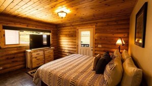 A log cabin bedroom with a bed, dresser, television, and a window. - Warm River Butte Lodge (Ashton)
