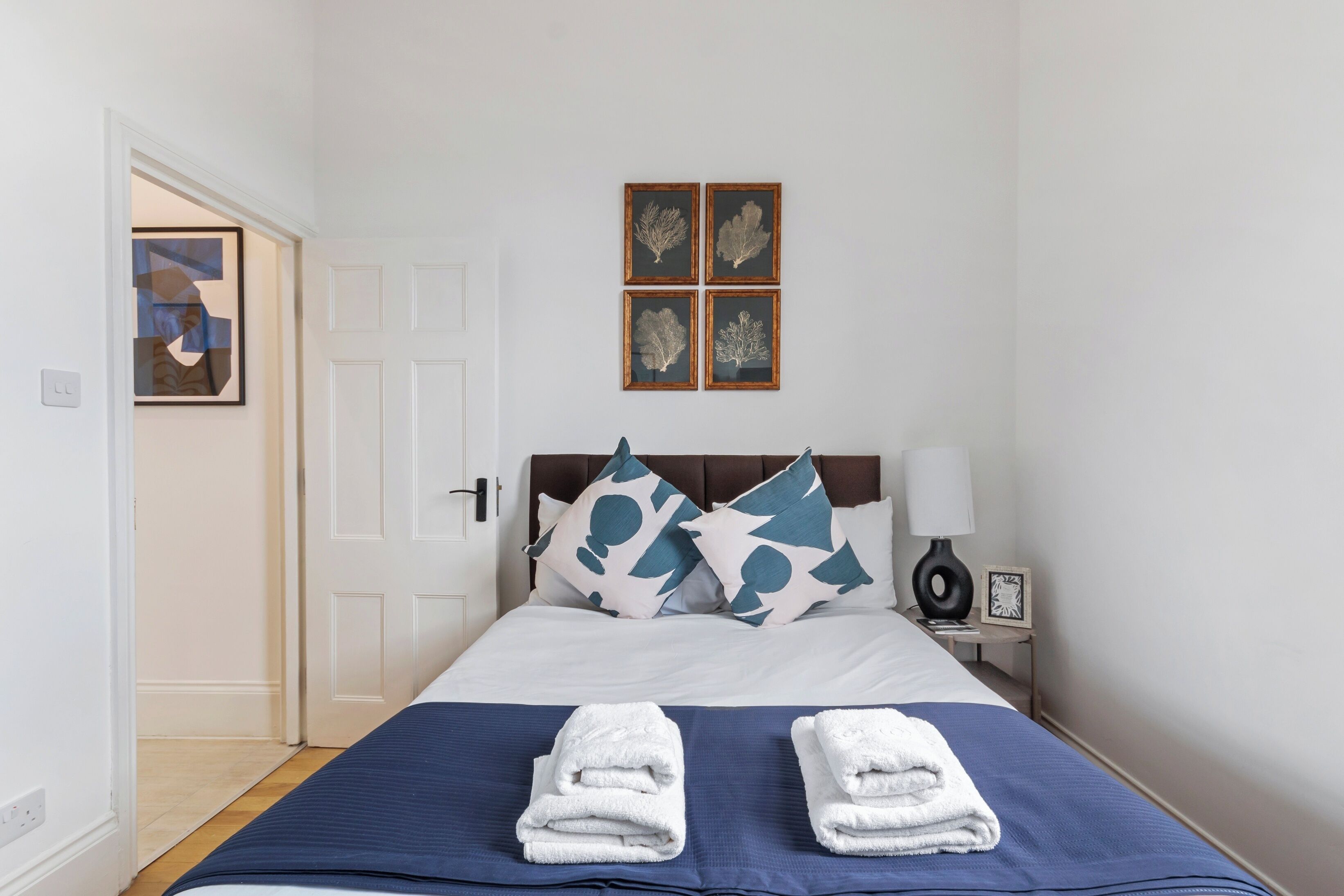 Un lit bien fait, recouvert d’une couvre-lit bleue, avec deux serviettes blanches pliées posées dessus. Quatre tableaux encadrés sont accrochés au mur, au-dessus du lit.