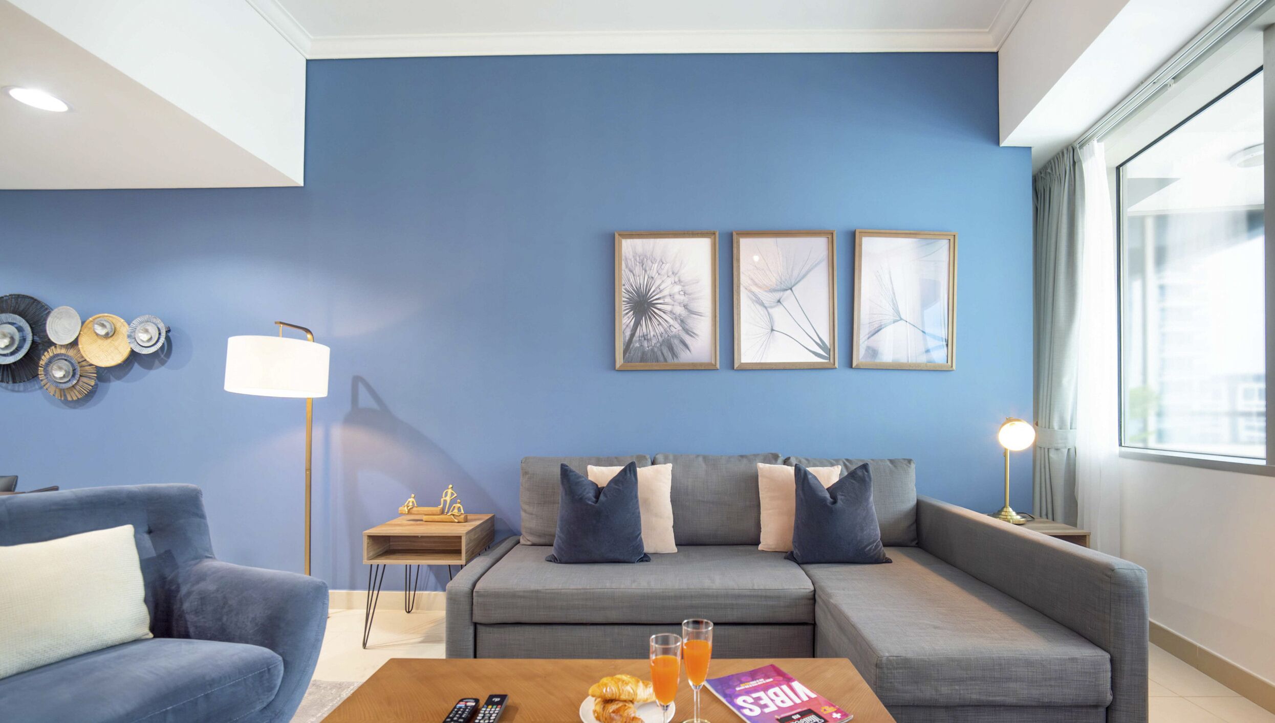 Un salon moderne avec un mur d’accent bleu, un canapé gris, une table basse avec des éléments pour le petit-déjeuner et des œuvres d’art encadrées.