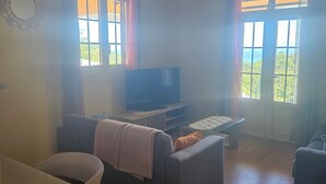 A living room with a sofa, a TV, and a table. There are windows with curtains and a view of greenery outside. - House between mountains and sea. (Deshaies)