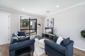 A modern living room with two sofas, a coffee table, a flat-screen TV, and a large window with a view of greenery.