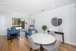 A modern living room with a round dining table, blue sofas, a TV, and a large mirror. - Grenache ~ Villa 1, Vineyard Views, Shared Pool & Wine Country (McLaren Vale)