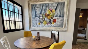 Un comedor con una mesa redonda de madera, dos sillas amarillas y una silla blanca. Un cuadro enmarcado con frutas y una vela sobre la mesa.
