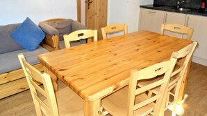 Een gezellige zithoek met een houten eettafel omringd door stoelen, een grijze bank met blauwe kussens en een keuken met een zwart aanrecht en kastjes.