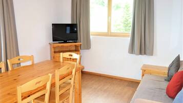 Una habitación con una mesa de comedor de madera, sillas, un televisor sobre un mueble de madera, un sofá con cojines y una ventana con cortinas.