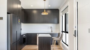 Une cuisine moderne avec des meubles de couleur sombre, un réfrigérateur en acier inoxydable, un îlot central doté de tabourets et un luminaire suspendu.