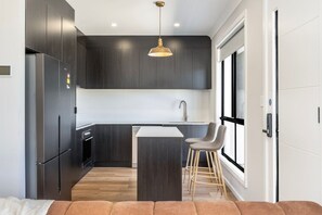 A modern kitchen with dark cabinetry, a stainless steel refrigerator, a central island with stools, and a pendant light.